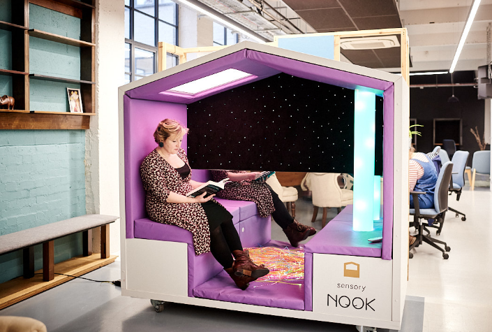 Woman sat in Nook Sensory Sanctuary in a workplace, reading a book and looking contented.