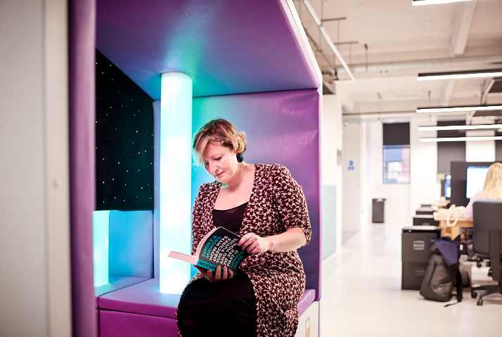 Woman sat in a Nook Sensory pod in an open plan workspace, looking cosy and relaxed reading a book.